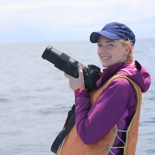 Frau mit Kamera und Schwimmweste, im Hintergrund das Meer