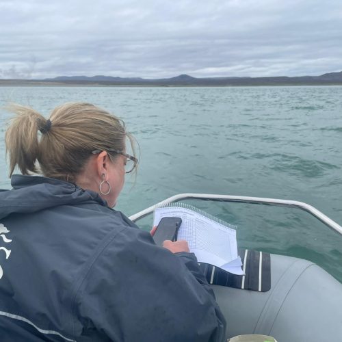 Frau auf Boot notiert Daten auf einem Blatt und nutzt gleichzeitig ihr Handy