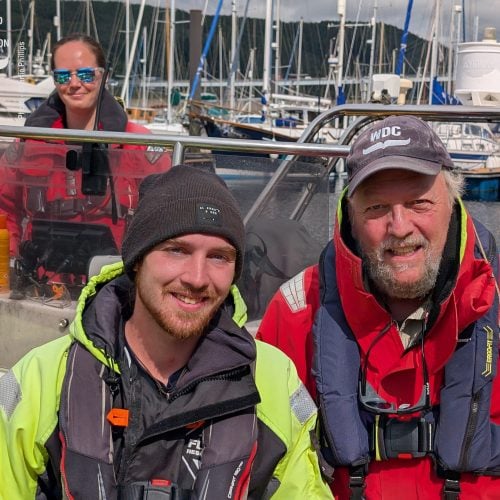 Ein jüngerer und ein älterer Mann in Overalls und Schwimmwesten am Hafen