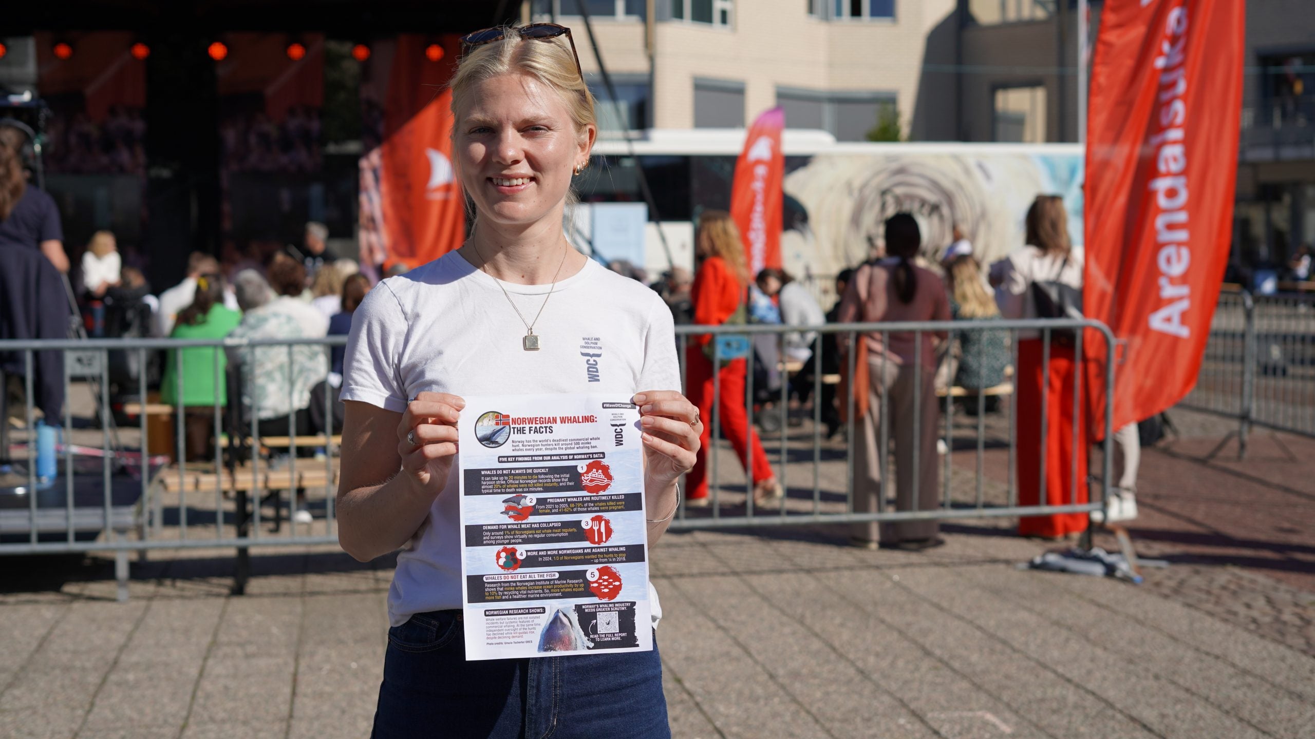 Frau im T-Shirt hält DIN-A4 Flyer hoch; im Hintergrund Fahnen und eine Bühne