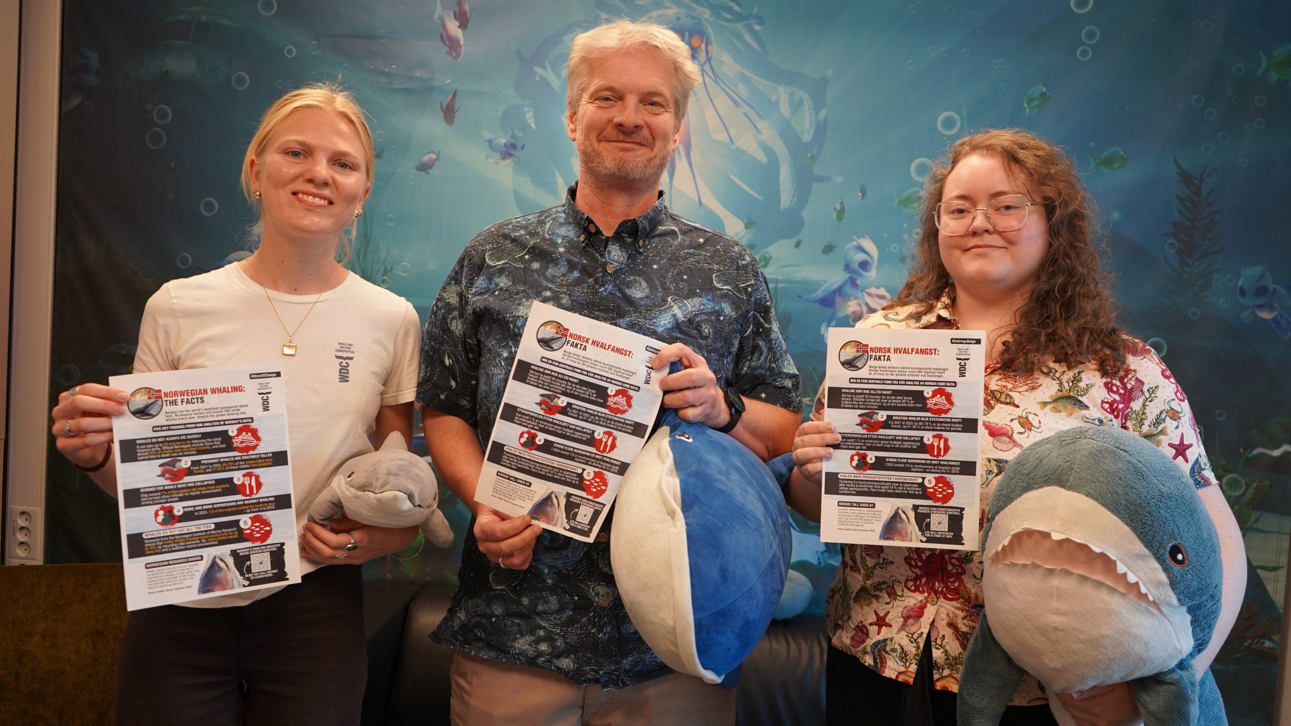 Drei Personen vor Unterwasserbild halten Infoblätter in die Kamera; zwei der Personen halten einen Plüschwal und einen Plüschhai fest
